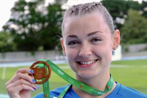 A person with braided hair and a blue shirt holds up a gold medal with a green ribbon, smiling. Trees and a track are visible in the background.