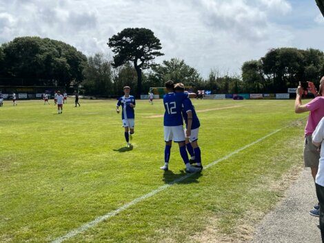 Three soccer players in blue jerseys, one with the number 12, celebrate near the sideline, while players from both teams are scattered around the field. Spectators watch from the sides on a partly cloudy day.