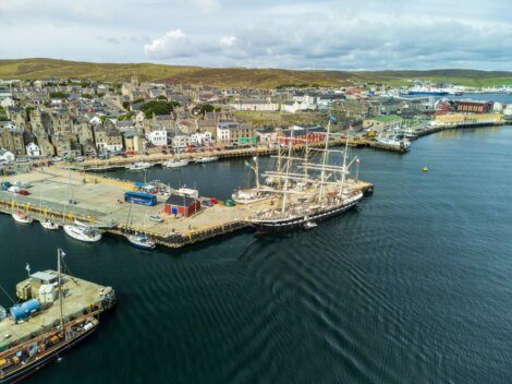 Aerial view of a coastal town featuring docks with boats and ships, colorful buildings, and rolling hills in the background.