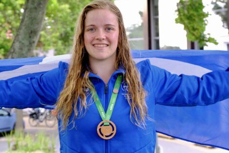 A person with long wet hair smiles while holding out a blue and white flag. They are wearing a blue jacket and a green lanyard with a bronze medal around their neck. Trees and a building are in the background.