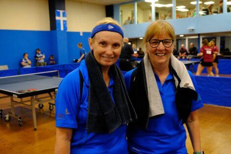 Two women in blue shirts with towels around their necks stand smiling in a table tennis facility. Other people and tables are visible in the background.