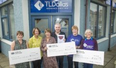 Five people stand holding large donation checks in front of a TL Dallas insurance office. The checks are made out to various charitable organizations.