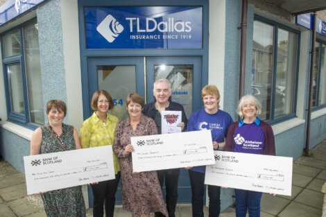 Five people stand holding large donation checks in front of a TL Dallas insurance office. The checks are made out to various charitable organizations.