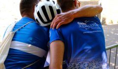 Three cyclists in blue jerseys stand next to a metal barrier, with one cyclist wearing a helmet and another placing an arm around a fellow cyclist’s shoulder.