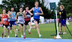 A group of male athletes running on a track, with one runner in a blue uniform leading the race. A woman is standing near the finish line, observing the race. Trees and spectators are in the background.