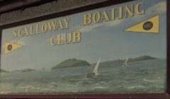 Signboard reading "SCALLOWAY BOATING CLUB" with sailing boats on the sea and islands in the background.