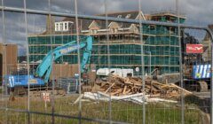 Construction site with a blue excavator, building material debris, and a partially completed green-clad building in the background, viewed through a chain-link fence on a cloudy day.