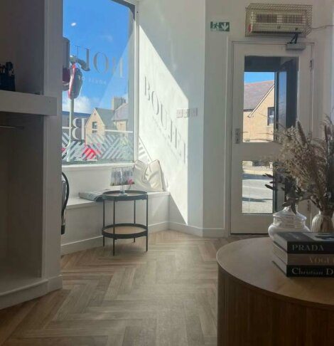 A cozy room with wooden flooring, a small round table with glasses, a door opening to an outdoor view, and a stack of books on a circular wooden table. Sunlight streams through the window.