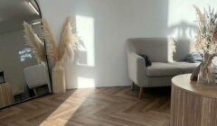 A minimalist room with herringbone wood flooring, a gray couch, a round wooden coffee table, a tall mirror, and decorative dried plants.