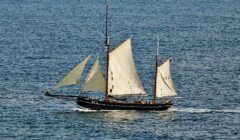 A large sailboat with white sails moves across the ocean, carrying several people on board. The water is calm, and the sky is clear.