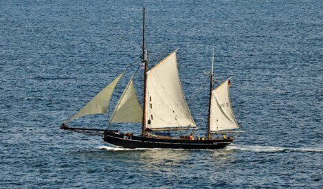 A large sailboat with white sails moves across the ocean, carrying several people on board. The water is calm, and the sky is clear.