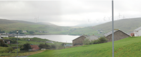 Hilly landscape with scattered houses and a lake, wind turbines on the hills in the background under a cloudy sky.