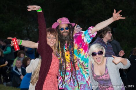 Three people posing outdoors, one in a colorful tie-dye outfit and hat, with others in casual attire. One holds a drink can, another gives a thumbs-up. Crowd and trees in the background.