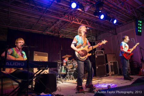 Four musicians wearing tie-dye shirts perform on stage with instruments including a keyboard, drum set, bass guitar, and electric guitar. Stage lighting and equipment are visible in the background.