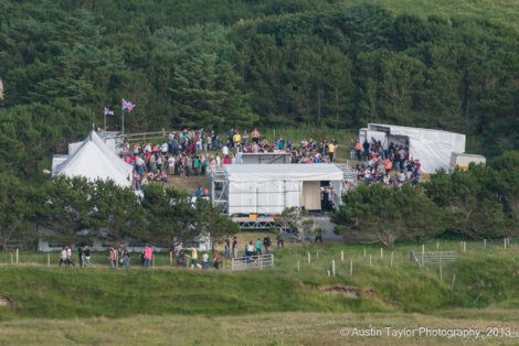 A crowd gathers around a tent and stage erected in a clearing surrounded by trees and grass. The setup includes white tents, various equipment, and a few flagpoles. Photo credit: Austin Taylor Photography, 2013.