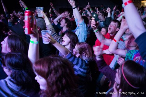 A crowd of people facing a stage with raised arms at a concert or event. Some are holding drinks, and many are wearing colorful wristbands. Photo credit: Austin Taylor Photography, 2013.