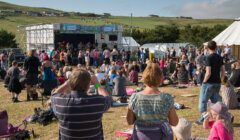 A crowd of people enjoying a live outdoor music performance at a festival in a rural area. Hills and some structures are visible in the background. The image is credited to Austin Taylor Photography, 2013.