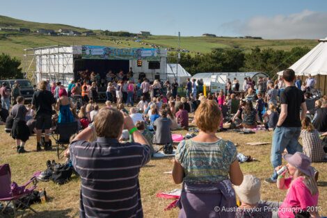 A crowd of people enjoying a live outdoor music performance at a festival in a rural area. Hills and some structures are visible in the background. The image is credited to Austin Taylor Photography, 2013.