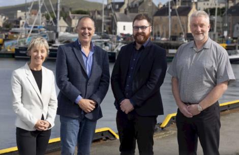 Four adults are standing together on a dock by a marina, with boats and buildings visible in the background.