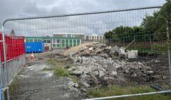 Construction site behind a metal fence with rubble and debris scattered on the ground. Blue containers are visible in the background, along with part of a building. Overcast sky above.