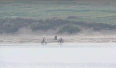 Three people ride bicycles along a sandy path by a body of water with grassy terrain in the background.