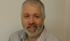 A man with gray hair and a beard, wearing a striped shirt, is smiling while sitting against a plain, light-colored wall.