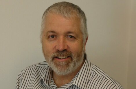 A man with gray hair and a beard, wearing a striped shirt, is smiling while sitting against a plain, light-colored wall.