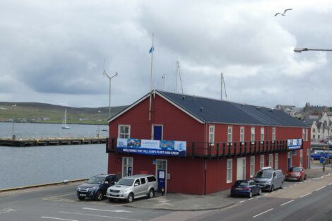 A red building labeled "Lerwick" with vehicles parked in front, situated next to a body of water with dock areas in the background and a seagull flying overhead.