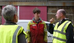 Three workers in high-visibility jackets engage in conversation outside an industrial building. One is gesturing with his hand while the others listen attentively.