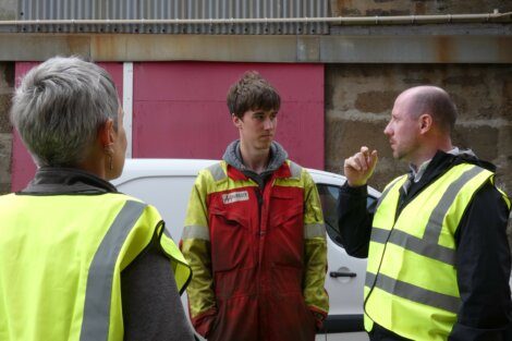 Three workers in high-visibility jackets engage in conversation outside an industrial building. One is gesturing with his hand while the others listen attentively.