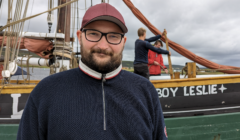 A person wearing glasses and a red cap stands in front of a boat named "Boy Leslie," where two other people are seen in the background handling the ropes.