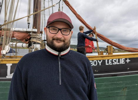 A person wearing glasses and a red cap stands in front of a boat named "Boy Leslie," where two other people are seen in the background handling the ropes.