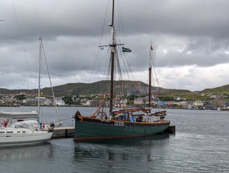 A large sailboat docked at a harbor with mountainous terrain and buildings in the background under an overcast sky.