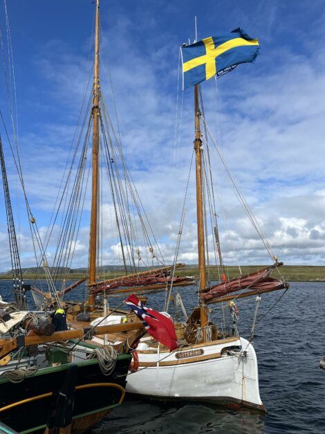 Two sailing boats docked at a marina, one flying the Swedish flag on its mast and the other displaying a red flag with a blue cross. The sky is partly cloudy.