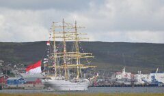 A tall ship with numerous sails and flags is docked in a harbor. The background features a hilled landscape and several buildings with a cloudy sky overhead.