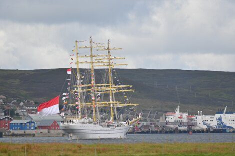 A tall ship with numerous sails and flags is docked in a harbor. The background features a hilled landscape and several buildings with a cloudy sky overhead.