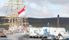 A tall ship with numerous flags displayed sails past a docked ferry featuring a blue graphic. Tugboats assist in the harbor. Hills and a smokestack appear in the background.