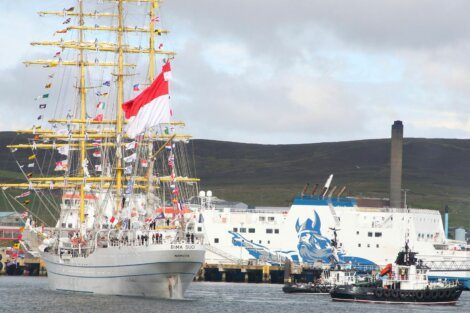 A tall ship with numerous flags displayed sails past a docked ferry featuring a blue graphic. Tugboats assist in the harbor. Hills and a smokestack appear in the background.