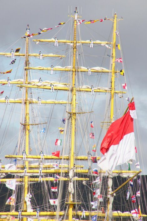 A tall ship with multiple masts and rigging is adorned with numerous colorful flags. A large red and white flag is prominently displayed on the right side of the ship. A cloudy sky forms the backdrop.