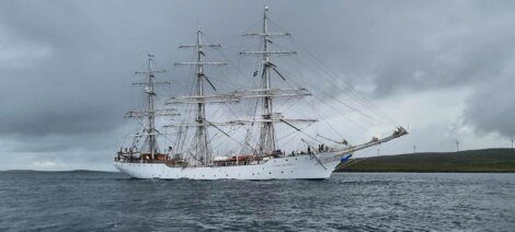 A large white sailing ship with three masts and numerous ropes sails on a body of water under a cloudy sky.