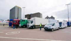 Several trucks and vans, including a caravan, are parked in an open area with a few people standing nearby. Tall buildings are visible in the background.