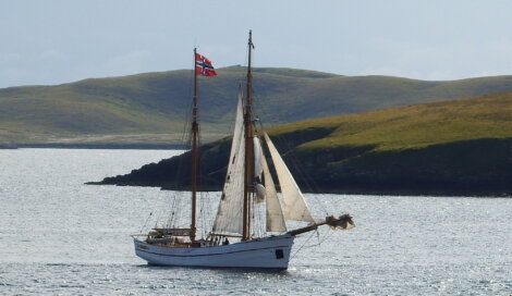 A two-masted sailboat with white sails and a Norwegian flag navigates through calm waters near hilly, green shores under a cloudy sky.