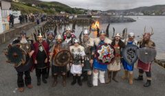 A group of people dressed in Viking costumes, holding shields and weapons, stands near a waterfront with a bonfire in the background and a crowd gathered on the pier.