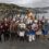 A group of people dressed in Viking costumes, holding shields and weapons, stands near a waterfront with a bonfire in the background and a crowd gathered on the pier.