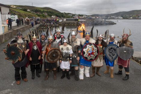 A group of people dressed in Viking costumes, holding shields and weapons, stands near a waterfront with a bonfire in the background and a crowd gathered on the pier.