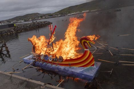 A model Viking ship with shields is set on fire on water near a rocky shore. Flames engulf the vessel, and partially burnt wood pieces float around it.