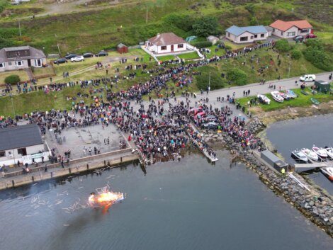 A large crowd gathers on a waterfront to watch a burning effigy floating in the water, surrounded by houses and boats.