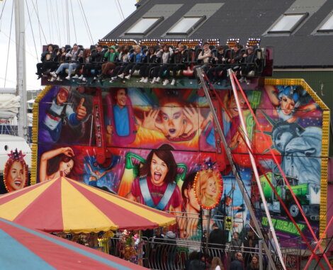 A colorful amusement ride with people seated at the top. The backdrop features vibrant, animated images of excited faces. Below, a striped tent and people walking are visible.