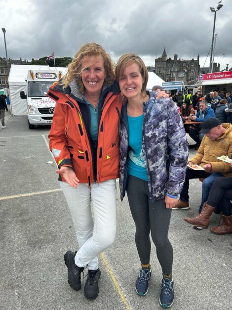 Two people in outerwear, one in an orange jacket and the other in blue, stand together smiling at an outdoor event with food trucks and people in the background.