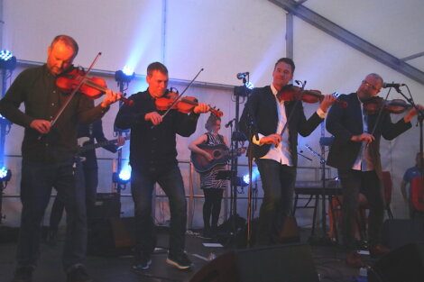 Four musicians playing violins on stage, a woman playing guitar, and a pianist in the background under a tent with blue stage lighting.